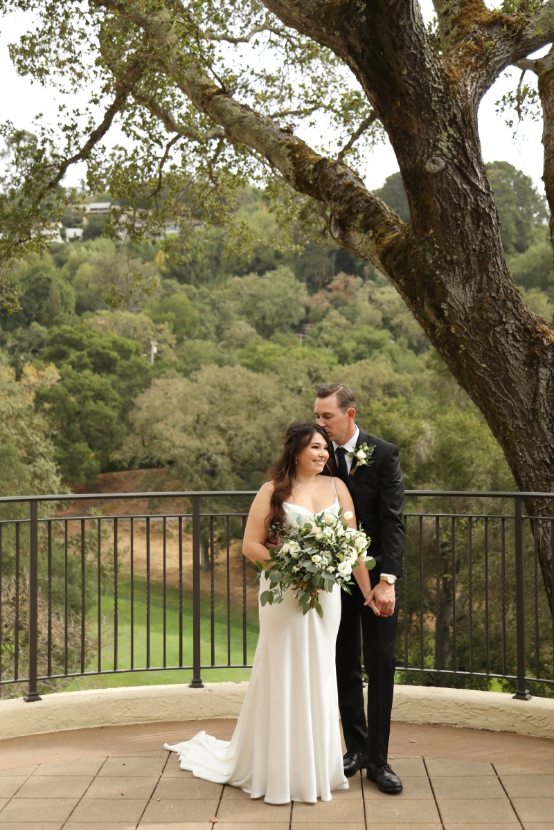 Will kisses Jane's temple on a terrace beneath an old oak, with the green Orinda hills behind them.