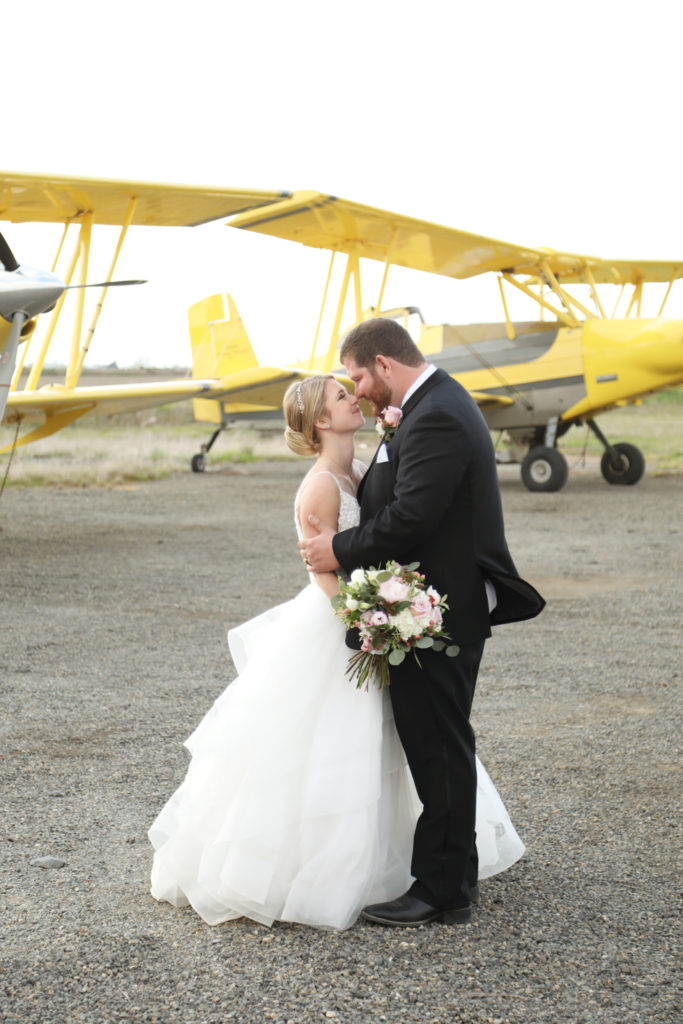 Nicole and Alex kiss on the airfield in front of yellow crop-duster biplanes.